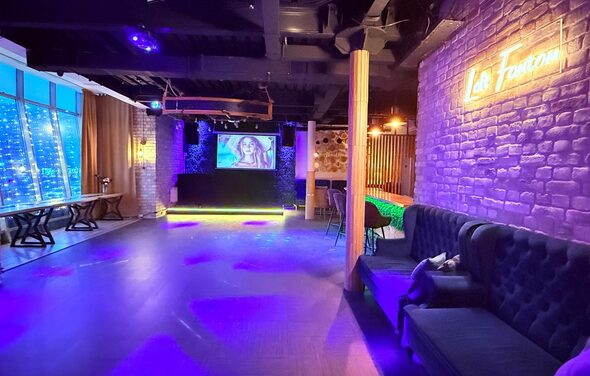 Лофт Фантом. Клубное пространство с деревянным полом. Левая стена — окна с тёмными шторами. Справа кирпичная стена с неоновым знаком. У дальней стены сцена с экраном. Мягкие диваны и стулья, неоновая и точечная подсветка.