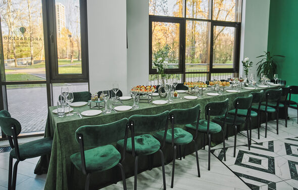 Arcobaleno Loft(Аркобалено Лофт). Пространство с длинным столом, покрытым зеленой скатертью, окружено стульями с зелеными бархатными сиденьями. На столе стеклянные бокалы, белые тарелки и закуски. Большие окна с видом на парк. Пол выложен узорчатой плиткой.