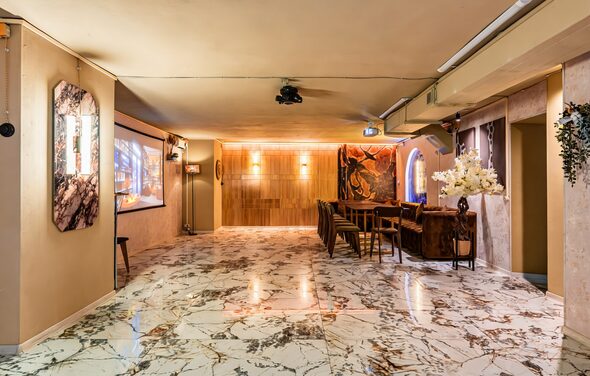 Стильный лофт Джаванди. Просторное помещение с мраморным полом, деревянной стеной, несколькими столами и стульями. На стенах висят картины и светильники. Потолочный проектор. Декоративные цветы и растения. Тёплое освещение.