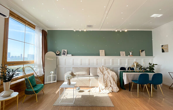 Зал Квартира. Уютное пространство в квартирном стиле. Гостиная с деревянным полом, зелёной акцентной стеной и белыми панелями. Диван с пледом, кофейный столик, обеденный стол с синими стульями, зеркало, растения. Большое окно с видом на город. Естественное и трековое освещение.