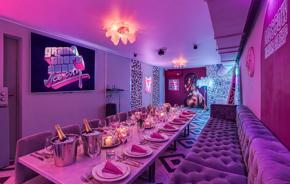 Неоновый лофт Гламур. Ресторан с длинным столом, сервированным бокалами и салфетками. Стены декорированы постерами и неоновыми вывесками. Мягкий диван вдоль стены. Потолочное освещение с лампами в виде цветов. Интерьер в розово-фиолетовых тонах.