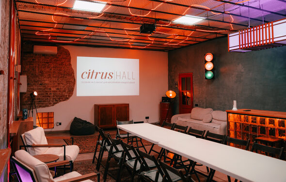 Citrus Hall (Цитрус Холл). Зал Бергамот. Лофт-пространство с кирпичными и серыми стенами. Длинный белый стол окружен черными стульями. На стене проектор с текстом. Нейтральное освещение с цветными лампами. Угловой диван, кресла, пуфы, светильники. Потолок с открытыми лампами.