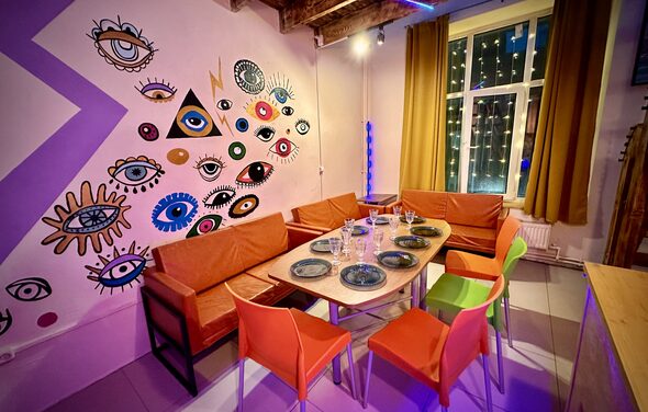 Яркий двухуровневый лофт. Стол с посудой. Яркие стулья с диванами. Обклеенные рисунками стена. Окно с навешенной гирляндой
