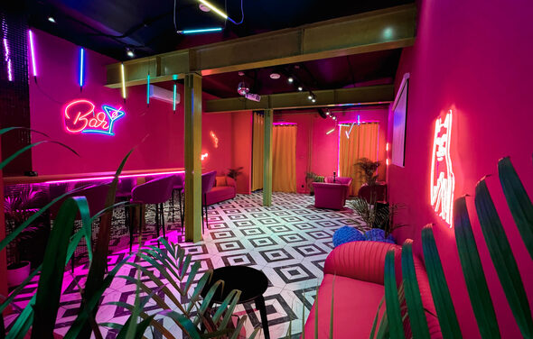 Плэйс Лофт. Prisma Loft(Призма Лофт). Барное пространство с розовыми стенами и неоновыми вывесками. Пол с черно-белой шахматной плиткой. Зона с барной стойкой и высокими стульями. Мягкая мебель, декоративные растения, яркое разноцветное освещение. Шторы тёплых оттенков.