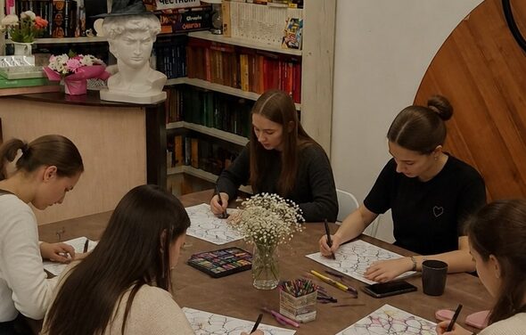 Творческая лофт-студия под мастер-классы и встречи. Комната с деревянными столами, на которых расположены цветные карандаши и блокноты. Несколько человек сидят за столом и рисуют. На заднем плане видны стеллажи с книгами, белый бюст и цветы в вазе. Светлое освещение, стены светлые.