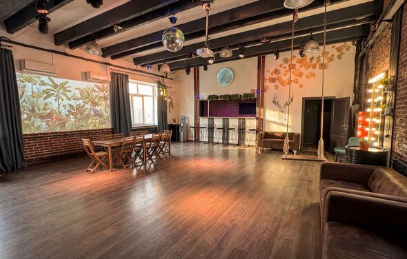 Лофт с верандой, мангалом и сценическим оборудованием. Большое помещение с деревянным полом и кирпичными стенами. Потолок с балками и подвесными светильниками. Барная стойка с высокими стульями. Диваны и обеденный стол с деревянными стульями. Декоративные элементы на стенах и зеркальный шар под потолком.