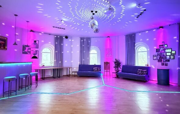 Центрум. Уютное светлое пространство круглой формы. Просторная комната с деревянным полом, мягкой мебелью в виде диванов, барной стойкой и стульями. Округлые окна с шторами. Декор дополняют растения и картины. Освещение разноцветное с акцентом на розовый, есть диско-шары. Стены белые.