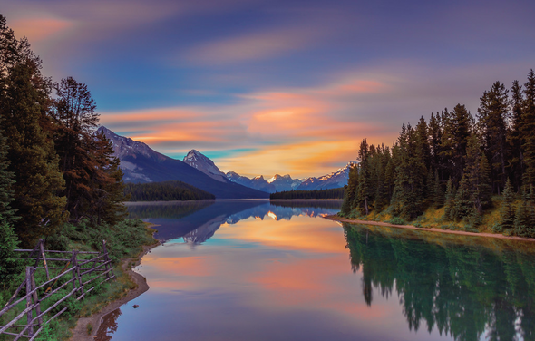 Тестовый зал мск. На картинке изображено живописное горное озеро, окруженное лесами и горами. Вода отражает красочное небо с облаками, окрашенное в розовые и оранжевые оттенки. На переднем плане виден деревянный забор и зелень.