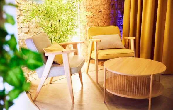 Большой лофт. Два кресла с подлокотниками, одно серое, другое желтое, деревянный круглый стол, кирпичная стена, желтые шторы, зеленое растение на фоне, мягкое освещение, светлые деревянные элементы, нейтральные и теплые тона помещений.