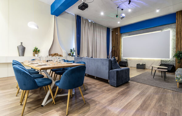 Rubin Loft (Рубин Лофт). Зал Сицилия. Современное праздничное пространство в светлых тонах с деревянным полом. Большой обеденный стол с синими стульями. Зона отдыха с серым диваном. Декоративное освещение, висящая лампа, проектор, занавески, декоративные элементы, растения.