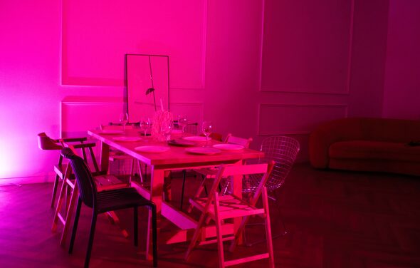 Стильный лофт для уютных вечеринок. Современное пространство с длинным деревянным столом, окруженным различными стульями, на столе посуда. Яркое розовое освещение. На стене панели, в углу диван. На столе зеркало. Пол паркетный.