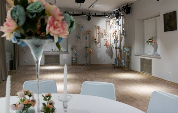 Пространство для ваших праздников. Просторное помещение с деревянным полом и белыми стенами. Круглый стол с белой скатертью и декором из цветов. На стенах и стойках декоративные цветочные композиции. Потолочные светильники, большое окно, приглушённое освещение.