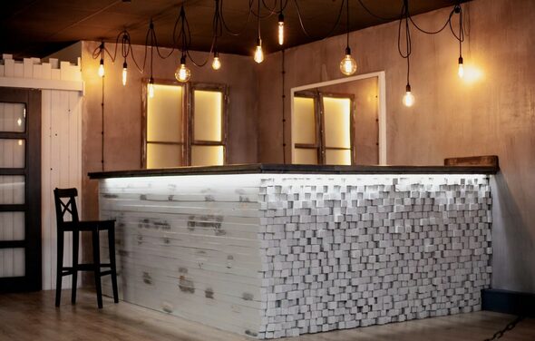 Лофт-пространство в Ярославле. Барная стойка с отделкой из белых деревянных блоков, стул, подвесные лампы с теплыми лампочками, деревянные стены с двумя окнами, пол светлого дерева, черный потолок с проводами, светодиодная подсветка стойки.
