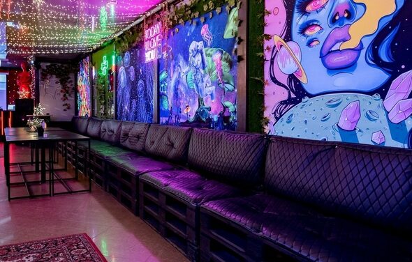 Яркий лофт для праздников. Праздничное пространство с длинным черным диваном и черными столами, яркие граффити на стенах, разноцветная светодиодная подсветка на потолке, ковёр с орнаментом на полу, стены украшены зелёными растениями, надпись "Social House" за диваном.