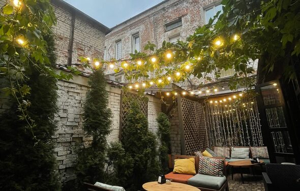 Лофт со своим двориком. Внутренний двор с деревянными столами и плетеными стульями с подушками. На стенах кирпич и зелень. Над пространством висят гирлянды с лампочками. В углу живая изгородь. На полу плитка. Стены окружающих зданий из кирпича.