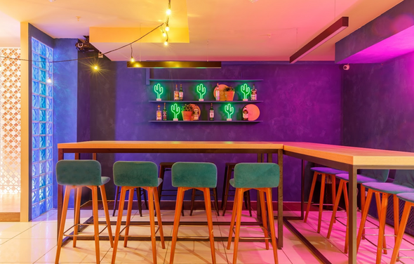 Антикафе с несколькими залами. Барная зона с высокими деревянными столами и барными стульями с бирюзовой обивкой. Темно-синие стены с неоновыми вывесками в виде кактусов. Подвесное освещение и настенная полка с декоративными элементами. Плиточный пол.