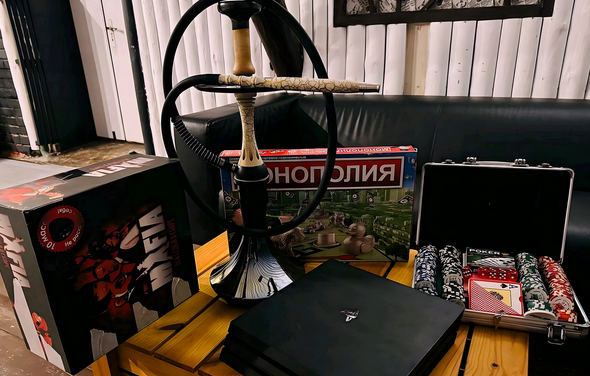 Лофт-мансарда . Кофейный столик из светлого дерева с кальяном, настольные игры «Мафия» и «Монополия», набор для покера с фишками и картами в открытом кейсе. На заднем плане черный кожаный диван и белая деревянная обшивка стен.