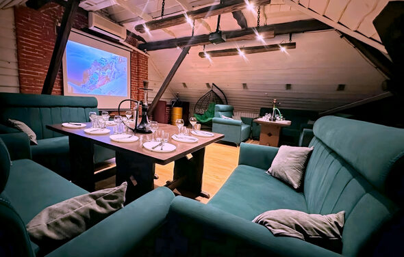 Лофт-мансарда . Лофт с деревянными балками, кирпичными стенами и наклонным потолком. Угловой диван и кресла бирюзового цвета, деревянный стол с сервировкой для ужина. Проектор с экраном, подвесные светильники, подвесное кресло. Светлое деревянное покрытие пола.