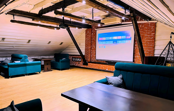 Лофт-мансарда . Лофт-пространство с деревянным полом, кирпичной стеной, наклонной белой крышей с деревянными балками и черными металлическими элементами. На стене экран с проекцией. Мебель: синие диваны и кресла, черный стол. Люстры с лампами на потолке.