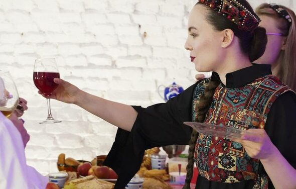 Лофт из белого кирпича в центре. Женщина в традиционной одежде держит бокал с напитком и тарелку. Белая кирпичная стена на заднем плане. На столе перед ней различные блюда и напитки. Теплое освещение.