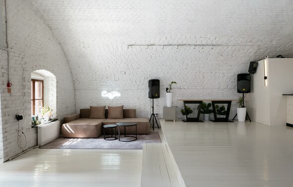 Brodniki Loft (Бродники Лофт). Лофт из белого кирпича. Арочное пространство с белыми кирпичными стенами и светлым деревянным полом. Коричневый диван с подушками на ковре, рядом с круглым столиком. По обеим сторонам украшения в виде растений в белых горшках. Два черных динамика. Окно с белыми рамами слева.