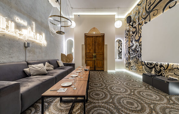 Rubin Loft (Рубин Лофт). Зал Дубай. Лофт-пространство с серым диваном, деревянным столом с тарелками и стаканами, подвесными светильниками, светлыми стенами с рисунками, деревянной дверью, неоновым освещением и декоративной печаткой на полу.