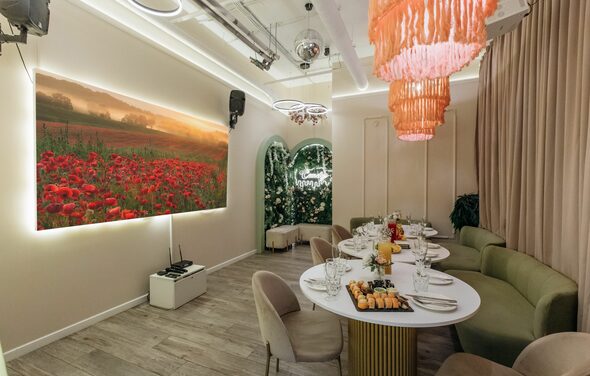 Лофт Кэнди. Пространство для мероприятий с круглым столом, сервированным посудой и закусками. Бежевые и зеленые кресла, светящиеся картины на стенах, яркие подвесные светильники. Светлые полы из дерева, драпировки на одной стене. На задней стене арка с белыми цветами.