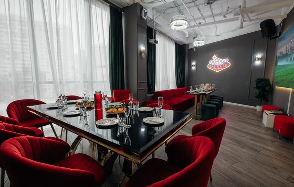 Rubin Loft (Рубин Лофт). Лофт Вегас. Современное помещение с застекленными окнами, красные кресла вокруг длинного стола с черной глянцевой поверхностью, сервировка столовых приборов, диваны у стены, неоновая надпись, люстры, деревянный пол, темные шторы, зеркало на стене.