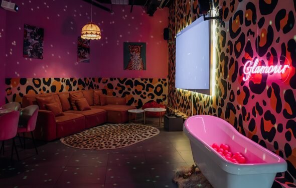 Лофт Гламур. Караоке-комната с леопардовым принтом на стенах, красный угловой диван, круглые стулья, столы, экран, неоновая надпись "Glamour", ванна с розовыми шарами, висячая лампа, коврик с узором. Теплое освещение, тёмные потолочные конструкции.