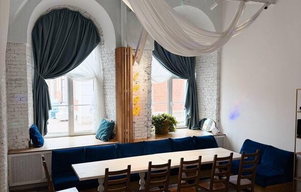 Малый светлый лофт для мероприятий. Помещение с высокими окнами и сводчатыми потолками. Белые кирпичные стены, длинный деревянный стол с деревянными стульями, мягкий синий диван. Шторы темно-синие, рядом кашпо с растением. На потолке ткани. Естественное освещение.