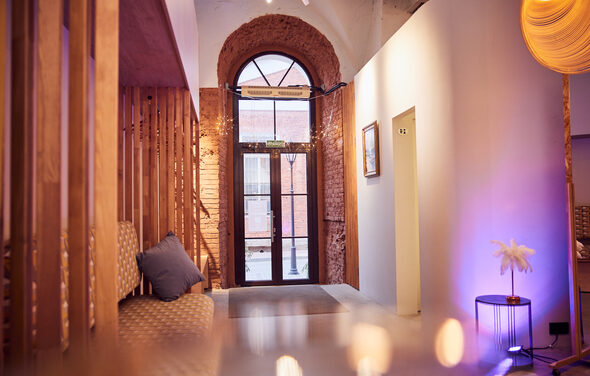 Малый светлый лофт для мероприятий. Арочное окно с деревянной рамой, стеклянная дверь, светлая стена с картиной, деревянная перегородка, мягкие стулья с подушками, фиолетовая подсветка, торшер, бетонный пол.