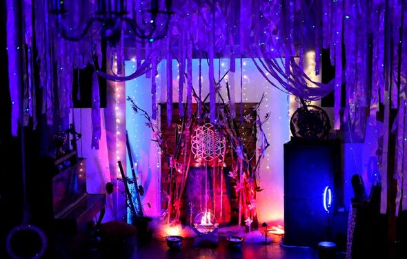 Лес. Театральный лофт с верандой, сценой и вторым ярусом. Темное пространство с ярким разноцветным освещением. Потолок украшен множеством висящих лент и гирлянд. В центре декоративная композиция с ветками и светящимися элементами. Видны мебель и оборудование, такие как фортепиано и аудиоколонки.