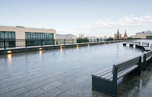 Площадка в Москве. Открытая терраса с брусчаткой, обрамленная перилами. По периметру установлены деревянные скамейки. Освещение вдоль ограждения. На фоне видны городские здания и собор. Светлый дневной свет.