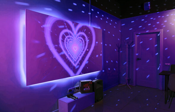Лофт-зал с караоке и диско-шаром. Комната с фиолетовой светодиодной подсветкой на стене, имеющей изображение сердца. На полу зеркало, рядом стоят видеопроектор и колонка. Справа дверь, в углу вешалка для одежды. Стены однотонные, потолок черный. Световые эффекты создают дискотечную атмосферу.