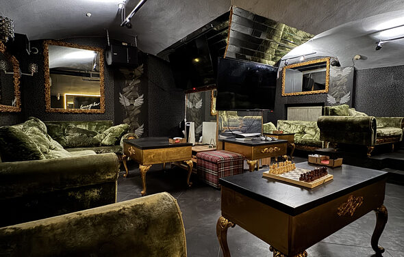 Shantaram(Шантарам). Лофт с темными стенами и потолком, зелеными бархатными диванами, золотистыми столами и рамами зеркал. На столах настольные игры и аксессуары. Есть крупный телевизор и освещение с потолка. Шахматная доска на переднем плане.