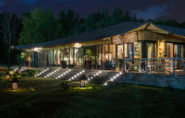 Soho Country Club (Сохо Кантри Клаб). Здание с большой террасой на фоне леса. Стеклянные стены, деревянные конструкции, крыша с вытянутым навесом. Перед входом ступени с боковой подсветкой. На газоне декоративные фонари и растительность. Внутри видны деревянные элементы и растения.