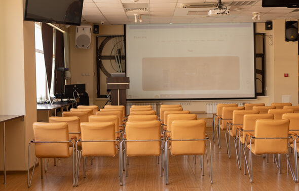 Зал 9. Просторный конференц-зал до 200 человек в Академии Кантаты. Конференц-зал с бежевыми стульями в ряд, деревянный пол, проектор с экраном, аудиосистема, кафедра, монитор, светлые стены, окна с занавесками, потолочные светильники.