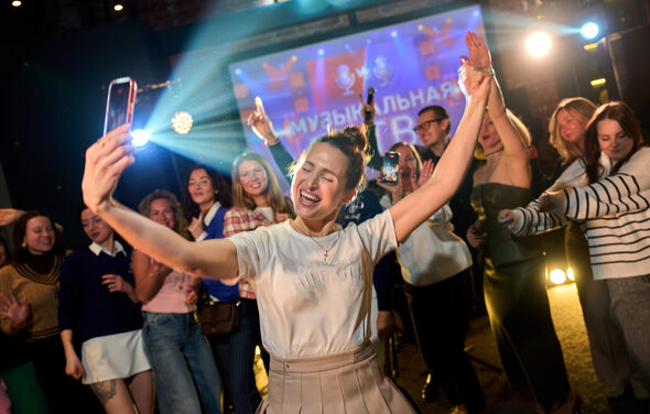 Лофт для мероприятий с гольф-симулятором. Люди танцуют перед проектором с текстом на экране, красочное свечение от прожекторов, множество молодых людей в неформальной одежде, светлые и темные тона в одежде, разнообразные эмоции и движения, вечернее освещение.