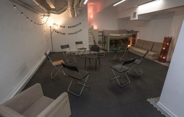 Пространство Нора лофт. Лофт-пространство с серыми стенами и потолком, украшенное гирляндами. По центру расставлены складные стулья, вдоль стены диван и кресло. Черный резиновый пол, лестница ведет на второй уровень. Надпись на стене из черных букв.