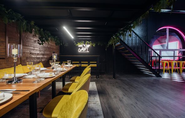 Rubin Loft (Рубин Лофт). Оушен Драйв. Просторное помещение с деревянным столом и желтыми стульями. Барная стойка с неоновым знаком. Стена отделана деревянными панелями, потолок черный, пол темный. Подсветка лампами и неоном. Поставлены декоративные свечи. Сбоку лестница с черными перилами.