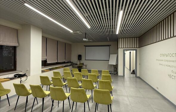 Зал 6. Просторный и светлый конференц-зал до 50 человек в Академии Кантаты. Конференц-зал с рядами желтых стульев, проектором и экраном. Светлые стены с текстом на русском языке, длинные занавески на окнах. Потолок с ламелями и встроенным освещением, пол серый, кафельный. В углу белая доска.