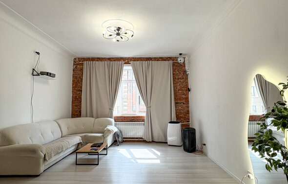 White Loft(Белый лофт). Уютный светлый лофт . Гостиная с белыми стенами, кирпичной акцентной стеной и большим окном с бежевыми шторами. Угловой белый диван, стеклянный журнальный столик, проектор на полке, колонка. Потолочный светильник, паркет светлого дерева, зеркало и растение.