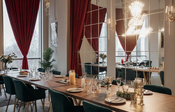 Зал Олд Мани. Лофт в индустриальном стиле. Просторное помещение с длинным деревянным столом, сервированным тарелками и бокалами. Тёмные стулья с мягкой обивкой. Зеркальная стена с подсветкой и надписью. Красные шторы, большие окна, декоративные растения. Мягкое освещение, светлые стены.