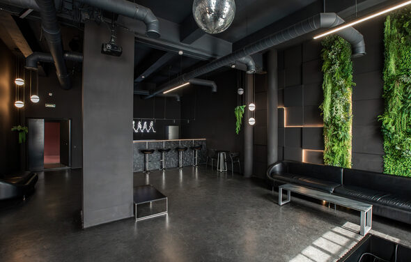 Rubin Loft (Рубин Лофт). Лофт Блэк. Современное помещение с тёмными стенами и полом, барной стойкой и высокими стульями. Вдоль стен чёрные кожаные диваны, столы из металла и стекла. Декоративные зелёные растения на стенах. Освещение с помощью подвесных и линейных светильников. Потолочные вентиляционные трубы.