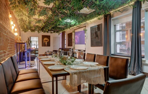 Лофт Семеновский. Праздничное помещение с длинным деревянным столом, окружённым кожаными стульями. На столе белые тарелки и бокалы. Кирпичная стена, занавешенные окна. Потолок украшен зеленью. Мягкое освещение. Окна с видом на город.