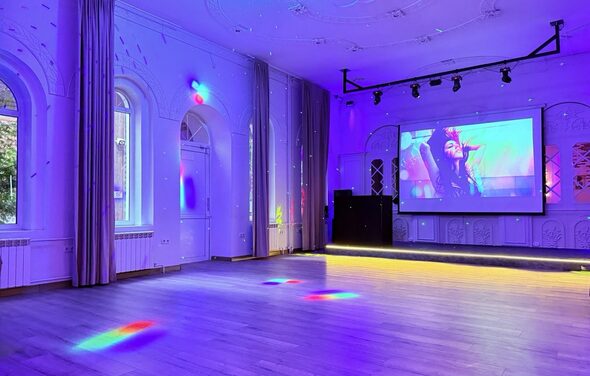 Лофт Софт. Просторное помещение со светлым паркетным полом и фиолетовой подсветкой. На стене большой экран с проектором, справа диджейский пульт. Окна с арочными рамами, закрытыми шторами. Потолочные светильники на треке и декоративные лепные элементы.
