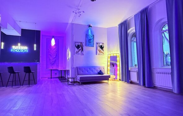 Лофт Лайф. Современное помещение с деревянным полом, высокой потолочной лампой и фиолетово-синим неоновым освещением. Зона с барной стойкой и стульями, диван, большие арочные окна с шторами, декоративные картины, высокое зеркало с подсветкой.