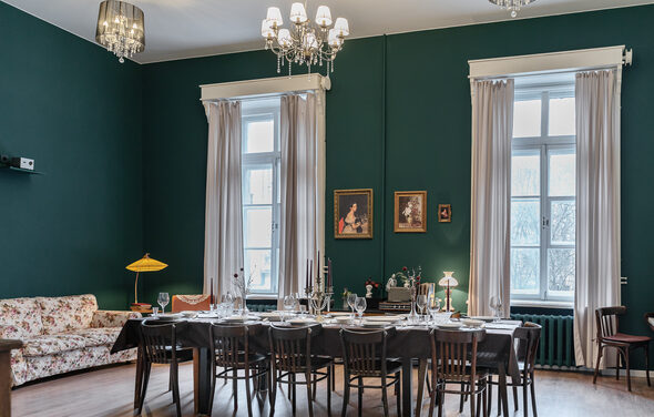 Пространство Имена. Винтажный лофт . Винтажная гостиная с зелеными стенами, двумя высокими окнами с белыми шторами. Просторный стол с черной скатертью, сервированный на двенадцать человек. Деревянные стулья, диван с цветочным узором. Люстры с лампами, картины на стене.