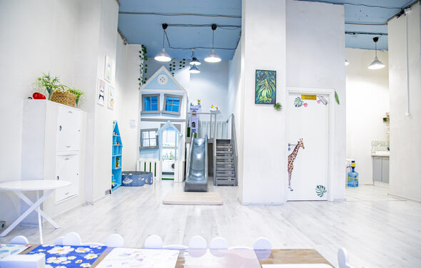 Лофт Облака. Зал Баунти на Полярной. Пространство детской игровой с зоной для занятий и горкой, большая белая комната с высоким потолком, белые стены и пол. Голубая игровая структура в виде домика с лестницей и горкой. Яркие акценты: жираф на двери, растения в горшках. Освещение подвесное.