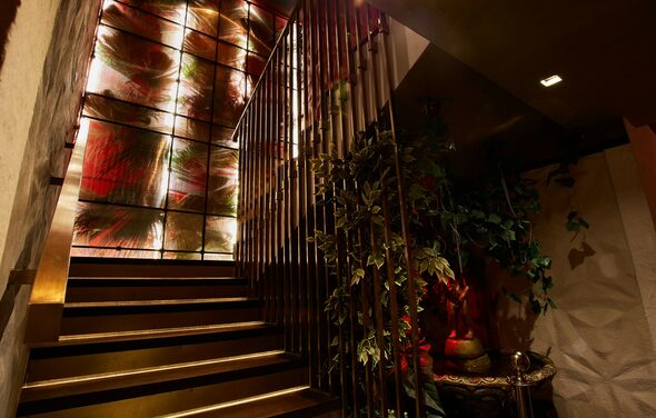 Микс Afterparty(После вечеринки). Лестница с металлическими перилами ведет вверх. Стена украшена панелями с абстрактным цветочным узором и подсветкой. Справа стоит декоративное растение в вазе. Освещение мягкое, коричнево-золотистые оттенки. Панели чередуют красные и зеленые цвета.